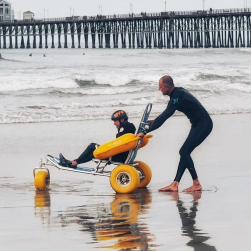 John wearing a wetsuit while pushing a person in a specialized beach wheelchair along the shoreline with a long wooden pier in the background.