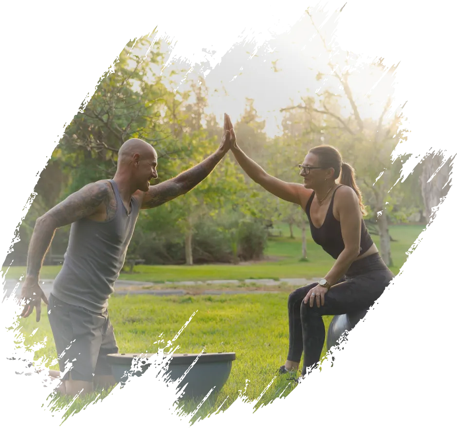 John and Christina exercising in a park, sharing a high-five while balancing on fitness equipment.