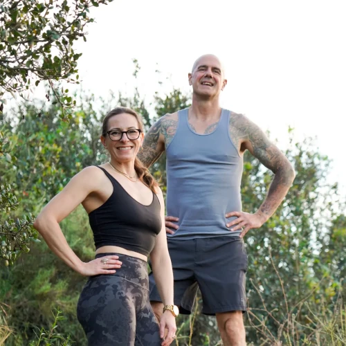 John and Christina posing together outdoors in athletic wear with green shrubbery in the background.