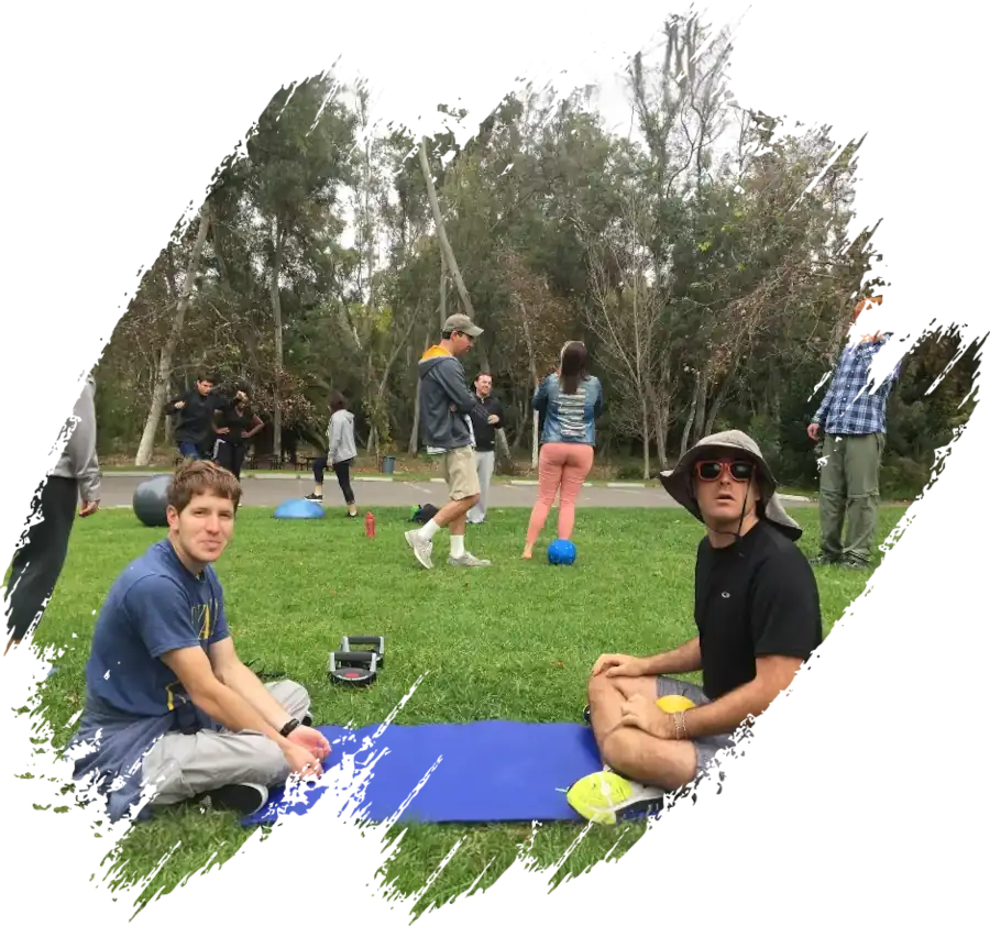 A group of people gathered in a grassy park area for an outdoor fitness session, with two men sitting on a blue yoga mat in the foreground.