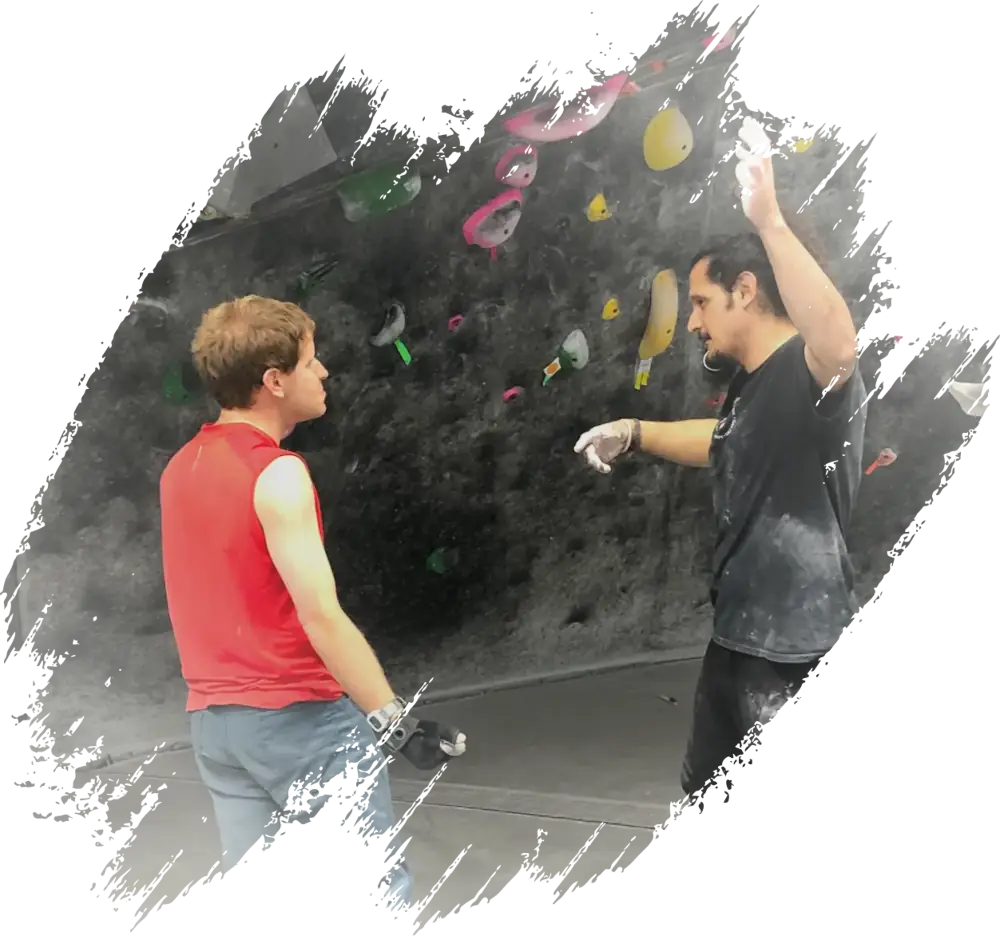 Two men talking in front of an indoor bouldering wall with colorful climbing holds.