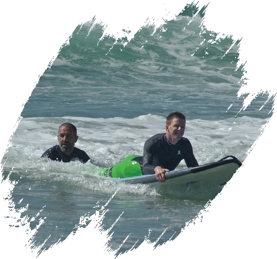 John stands in the ocean waves, guiding a man who is lying prone on a surfboard.