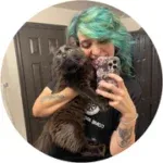 A mirror selfie of a smiling woman with bright green and blue hair holding a large grey cat.