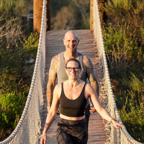 John and Christina smiling as they walk across a wooden suspension bridge with rope railings.